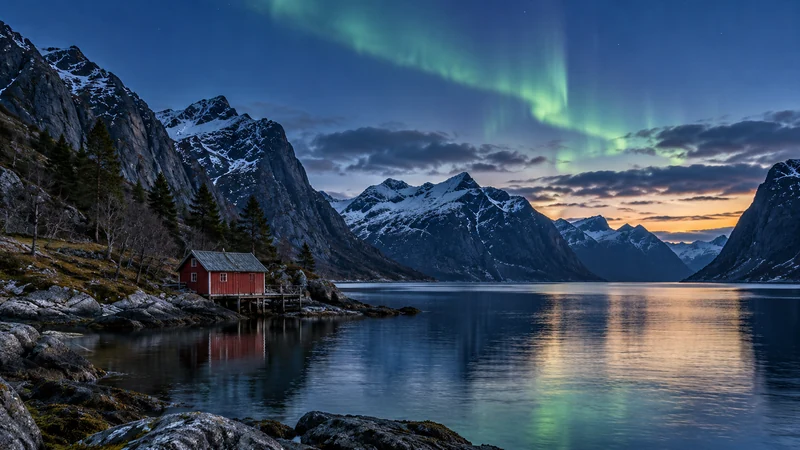 Nordlys over en fjord med snødekte fjell og et rødt naust i forgrunnen.